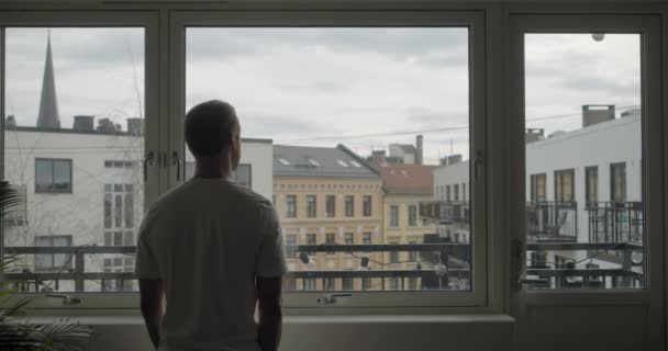 ノルウェーの若者だけを撮影し 都市の背景を持つ暗いアパートから窓の外を見てコロナウイルス検疫で隔離された広い静的バックシネマカメラノルウェーのオスロの中央部 — ストック動画