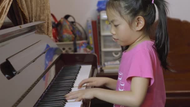 自宅でピアノを弾くアジア系の少女のサイドビュー ピアノのレッスンを練習 7歳の子供の女の子 長い黒い髪 髪のネクタイ — ストック動画