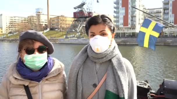 Švédsko, Stockholm. Portrét matky a dcery, jak se dívají do kamery a nosí ochrannou masku proti znečištění a koronavirové pohotovosti. Švédská vlajka mávající v pozadí