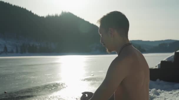 一个没有衬衫的人坐在冰封的山湖边沉思 地面上结着雪 年轻人准备好在冰冷的冰水中进行极端的冒险和游泳 — 图库视频影像