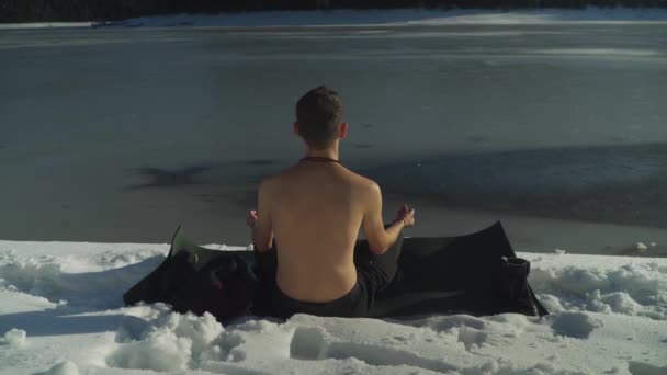 Muž bez košile, který sedí na černé podložce jógy ve sněhu vedle zamrzlého jezera a medituje. Mladý muž těší frigidní arktické podmínky v teplém slunečním světle koncentruje a medituje pokojně.