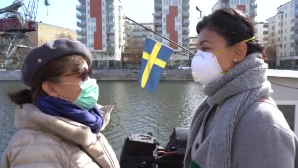 Portrét asijské matky a dcery, jak se dívají jeden na druhého a nosí ochrannou masku proti koronaviru. Modré moře a švédská vlajka v pozadí. Virová ochrana, Švédsko