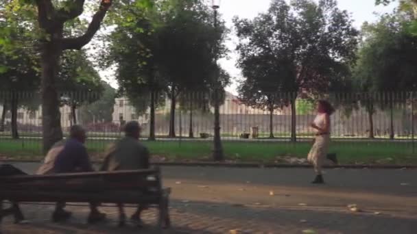 若い女性が並木道を走り2人の先輩がベンチで彼女を見ている — ストック動画