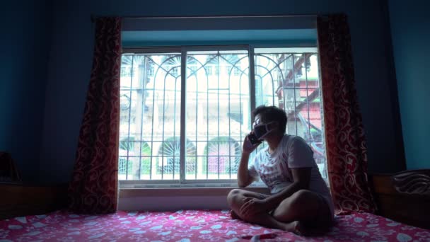 由于Covid Coronavirus Lockdown 自我隔离和社会疏离 亚洲年轻男子在靠近窗户的家里通过电话交谈 — 图库视频影像