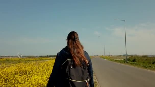 A video of following a woman walking at the street full of yellow mustard flowers.