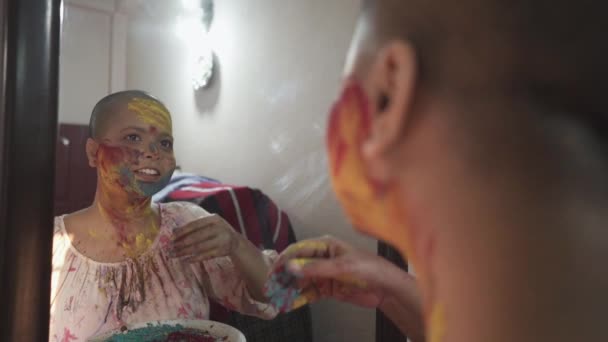 希望の癌患者のはげの女の子は 色のホリ インドの祭りの鏡の前に自宅で頬に色の粉を適用します — ストック動画