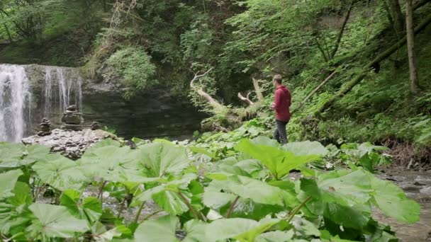 Fotograf v červeném svetru se prochází a snaží se najít kompoziton pro vodopád. Fotografie v přírodě fotografující vodopád s fotoaparátem Fujifilm a stativem.