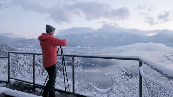 湖の上の日の出をキャプチャするために三脚に富士フイルムカメラを搭載した写真家が出血した 日の出に雪の中を歩く写真家 Bled Lake — ストック動画