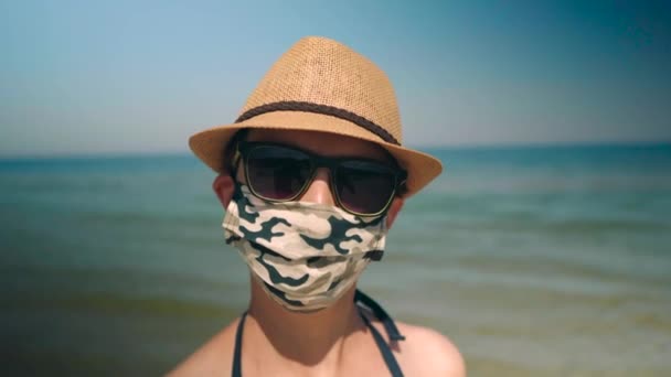 Žena nosí koronu obličejovou masku se slunečními brýlemi a kloboukem na pláži před oceánem