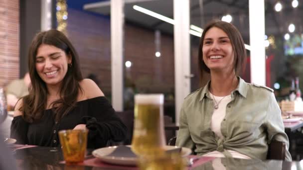 Dvě krásné brunetky dívky se usmívají a baví v restauraci s přáteli. Užívám si chvíle, povídání a povídání u večeře.