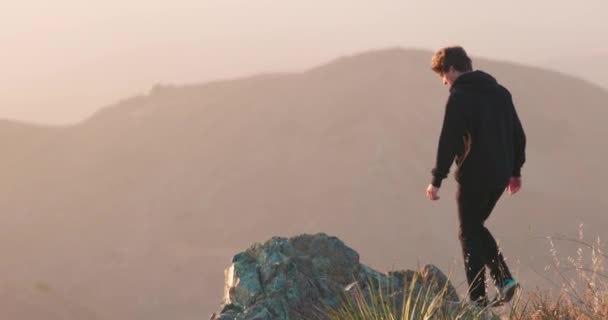 若い大人は日没時に霧の多いカリフォルニア渓谷を見下ろす険しい山の縁に出て行きます — ストック動画