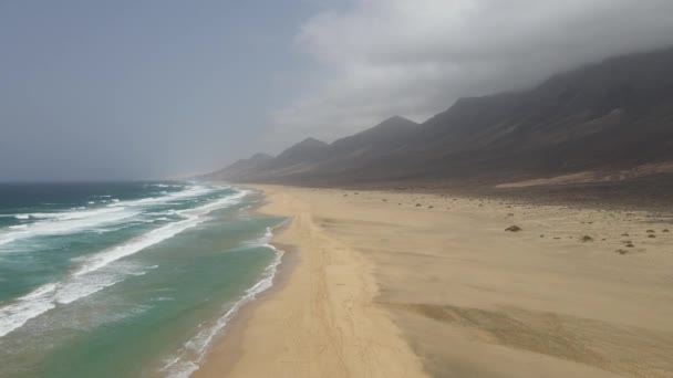 カナリア島フエルテベントゥラ島を背景に美しい山々とコーヒーの長い白いビーチに墜落した大きな波の空中ドローンショット — ストック動画