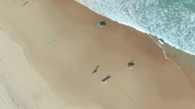 Sörfçülerin hava görüntüsü. Küçük dalgalar ve derin mavi deniz manzaralı Atlantik Okyanusu 'nda güneşli bir gün. Sahilde sörf tahtası tutan ve yürüyen sörfçülerin olduğu bir grup.