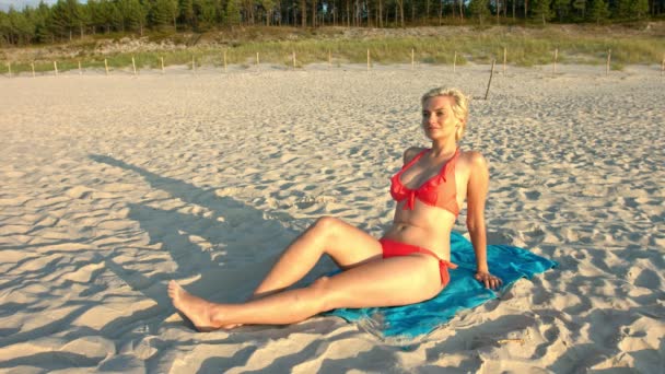 一个性感的女人坐在沙滩上 为一个完美的静态照片姿势 一位穿着时髦比基尼的优雅女士在阳光下独自坐着 脸上带着轻松的表情 一个悠闲的女人独自笑 — 图库视频影像