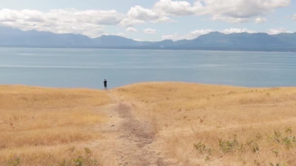 美しい黄金の牧草地とカナダのブリティッシュコロンビア州ホーンビー島の山々で海を見下ろすヘリウェル草原の端に立つ男 — ストック動画