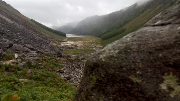 曇りや雨の日にアイルランドの場合は岩の多い山の中でハイカーの登山のショットを明らかに 背景に自然湖と美しい丘の風景のパンショット アイルランド ヨーロッパ — ストック動画