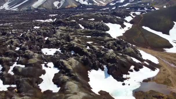 アイスランドのLandmannalauarの虹の山の渓谷に白い雪のパッチを持つ黒い溶岩フィールド ハイキングコースの上の空中 前方に押してカメラの動きを傾ける — ストック動画