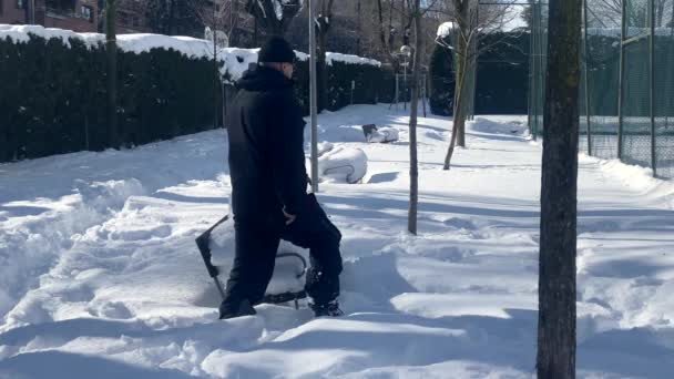 スノースーツを着た男とサングラスは雪の公園を歩く 雪は公共のベンチや地面をカバーしています 彼は雪に覆われたベンチに座っている — ストック動画