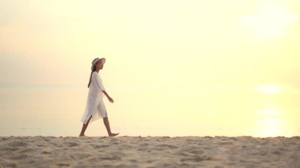 一个穿着宽松外套的漂亮女人在日落时分沿着海滩散步 — 图库视频影像