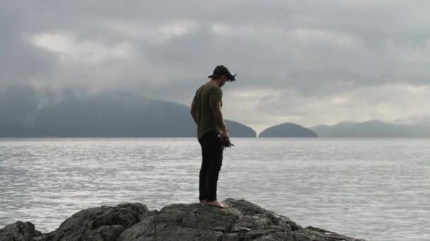 Fpvクワッドコプターを持つ白人男性は オーバーキャストで穏やかな海を見下ろす岩の上に立っています — ストック動画