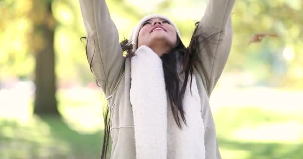 Attractive Girl tossing colorful autumn leaves up in air in park. The foliage falls down over herself.  She is wearing a white jacket and beanie on a chilly fall day. Shot in 4k slow motion.