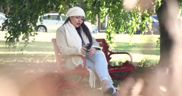 寒冷的秋秋季节 妇女们坐在城市公园的长椅上发短信 她停了下来 环顾四周 穿着一件白色的夹克和一件便帽 在4K内开枪 — 图库视频影像