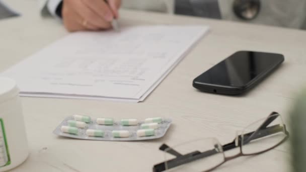 Orvos ír a fehér könyvben a szemüvegével, a mobiljával és a kapszula gyógyszerével az asztalnál.. 