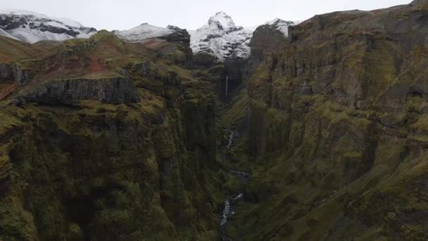 从空中俯瞰冰岛北冰洋的深谷 无人机在峡谷内飞行 狭窄的溪流在峡谷内飞行 背景是雪山 — 图库视频影像
