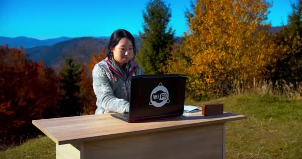 山の中のテーブルの後ろのノートパソコンで仕事をしている日本人女性 背景には秋の森 リモートワーク 自宅からの仕事 遊牧生活 フリーランスのライフスタイル 研究コンセプト — ストック動画
