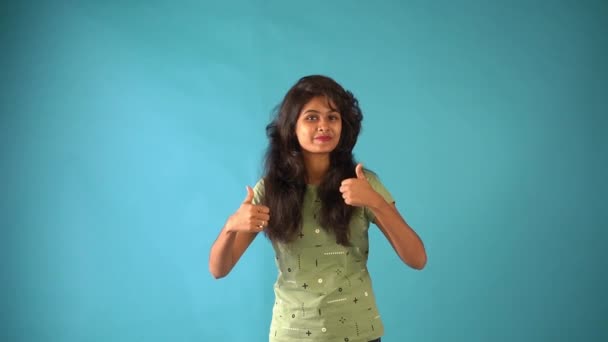 孤立した青の背景スタジオに立っているカメラを見て親指アップを示す緑のTシャツの若いインドの女の子 — ストック動画