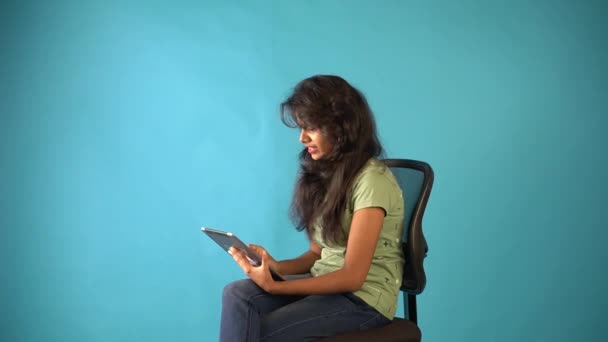 一位身穿绿色T恤衫的印度小女孩坐在一个偏僻的蓝色背景工作室的椅子上 一边在视频聊天一边聊天 — 图库视频影像