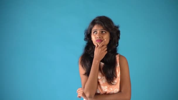 Ein junges indisches Mädchen in orangefarbenem Gewand, das besorgt in die Kamera blickt und dann in einem isolierten blauen Hintergrund denkt.