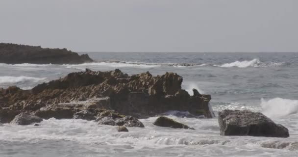 曇りの日にビーチの岩の海辺に波がぶつかったり飛び散ったり — ストック動画