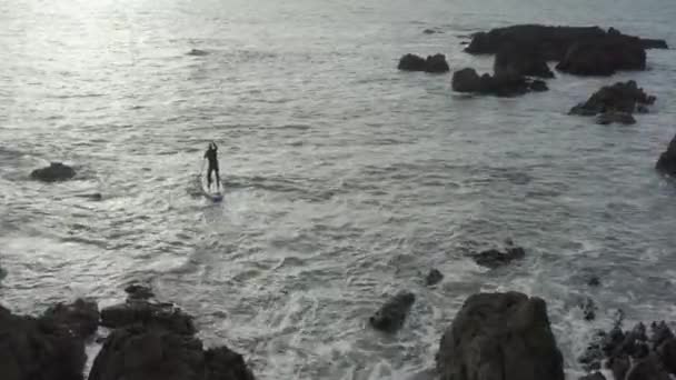 Aerial Följer Hanen Paddla Boarder Hackig Ocean Nära Skarpa Stenar — Stockvideo