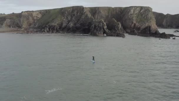 青いパドルボード上のウェットスーツの男険しい海の崖に向かってパドル — ストック動画