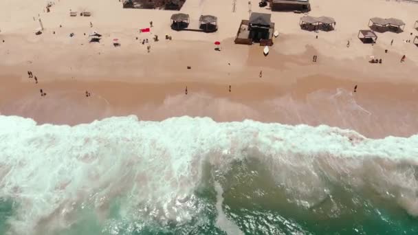 砂浜の海岸線で波が打ち寄せる無人偵察機です人々が海岸にぶら下がっている間にライフガードステーションが真ん中にあります — ストック動画