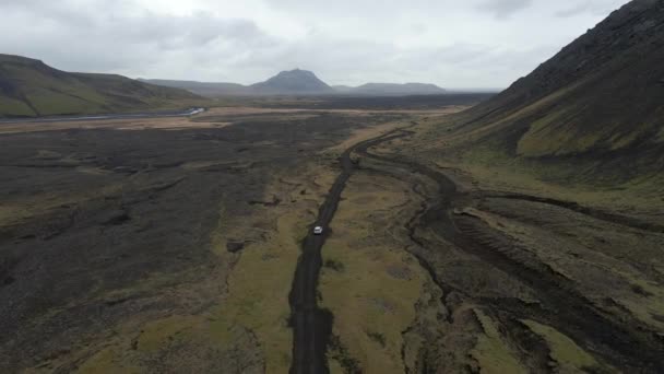 曇った日にアイスランドの両側に黒い砂と山々と道路に沿って運転する白い車を追跡するドローン — ストック動画