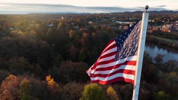 秋の黄金の時間の日の出の光の間に風に振ってアメリカの国旗の映画的な航空 カラフルな葉 アメリカ美しく誇りに思うアメリカ 町は川に映る — ストック動画