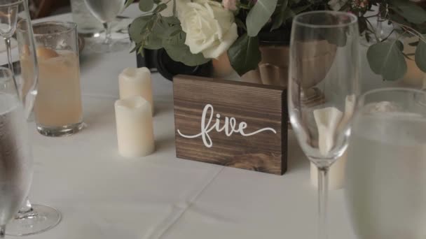 Wedding dining with a view of wooden decorations surrounded by candles, flowers and tall glasses. Sign reads Five in cursive