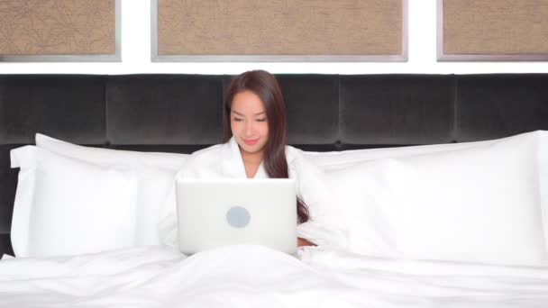 Working from the comfort of a big comfy bed, a young attractive woman in a bathrobe scans her laptop as she prepares for work.