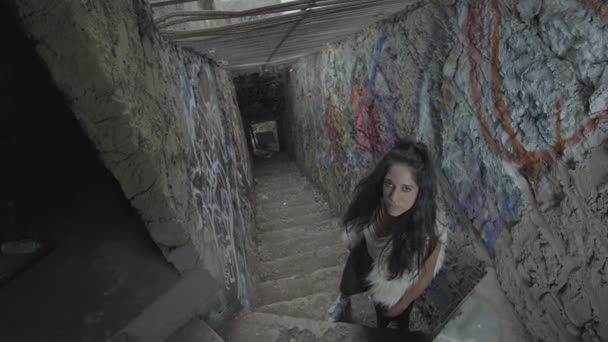 一个眼神凝视着的年轻女人站在楼梯顶上 在一条狭窄的走廊里 上面涂满了涂鸦 天花板上还挂着铁棍 — 图库视频影像
