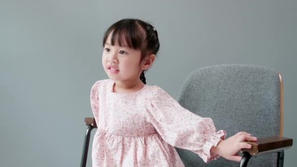 3岁的快乐快乐的亚洲孩子坐在灰色背景前的椅子上 — 图库视频影像