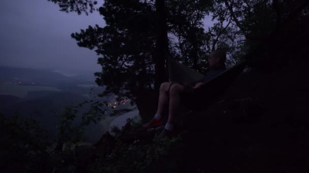 一个穿短裤的白人坐在山顶上的吊床上 现在是晚上了背景是一条河 村庄的灯光朦胧 还有一片云雾弥漫的树林 — 图库视频影像