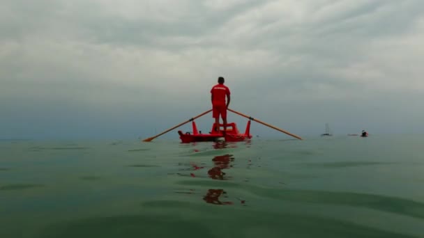 赤いイタリアのライフガードは カヤックで人々を見ている間 緊急時の待機とボートの救助ボートを漕ぐ準備ができています 低角度海水面 — ストック動画