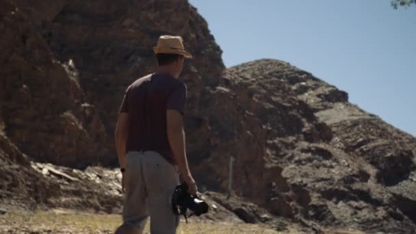 背景に山があるアフリカの河床をカメラが歩くスタイリッシュな帽子をかぶった写真家の手持ち撮影 — ストック動画