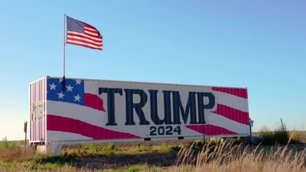 ドナルド トランプ2024大統領選挙運動アメリカ国旗が風に吹かれて署名する どこの真ん中にも背の高い草を持つ農村農場の土地の大型トラックやトレーラーに描かれた — ストック動画