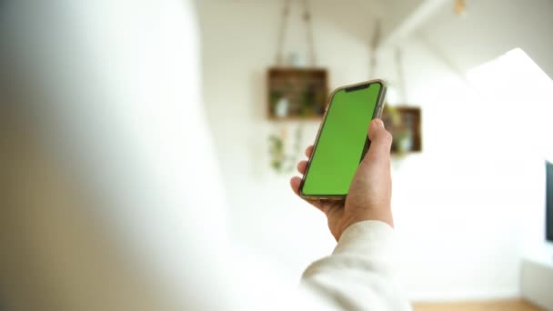 Egy férfi kezében egy okostelefon zöld képernyő függőleges fehér otthoni háttér. Koncepció alkalmazáshoz, online vásárláshoz vagy lekérhető videóhoz.