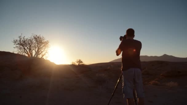 Dramatický široký statický snímek turistického fotografa, jak fotografuje krajinu směrem ke slunci na stativu v poušti v Africe.