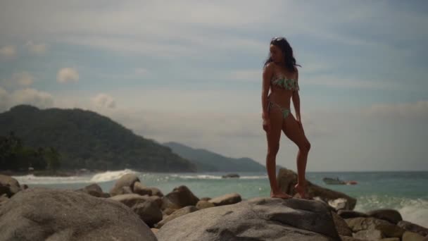 Gyönyörű fiatal nő bikiniben áll egy sziget sziklás partján, a hullámok lassított felvételben hullanak.