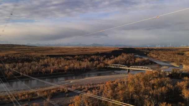 カナダのアルバータ州南部の送電線を後方に飛行する無人機 電柱に接近中 ロッキー山脈の郊外にある美しい古い橋の映像 — ストック動画